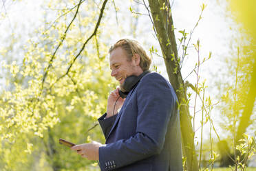 Lächelnder Geschäftsmann, der ein Smartphone benutzt, während er im Park an einem sonnigen Tag steht - JOSEF01474