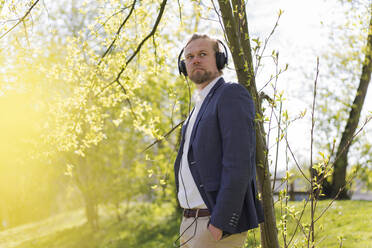 Männlicher Unternehmer hört Musik, während er an einem sonnigen Tag in den Park blickt - JOSEF01476