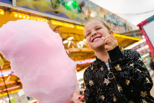Von unten erfreutes Mädchen, das lächelt und süße Zuckerwatte isst, während es auf dem Rummelplatz steht - ADSF10184