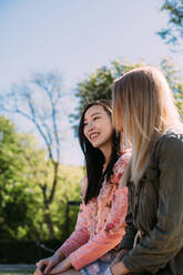 Two multiracial young women in casual outfits talking and looking at each other while sitting on bench in park - ADSF10187