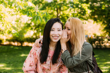 Junge kaukasische Frau lächelt und flüstert Geheimnis ins Ohr des lächelnden asiatischen Freundes, während sie Zeit im Park zusammen verbringen - ADSF10188