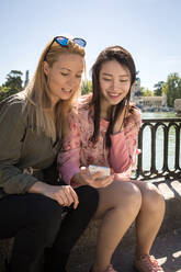 Junge multirassische Frauen in trendigen Outfits lächelnd und Surfen Smartphone beim Sitzen in der Nähe Böschung Geländer auf sonnigen Tag in der Stadt Straße - ADSF10190