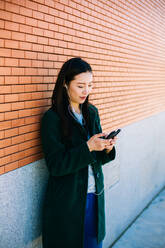 Young Asian woman listening to music and browsing smartphone while leaning on brick wall on city street - ADSF10191