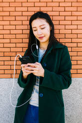 Young Asian woman listening to music and browsing smartphone while leaning on brick wall on city street - ADSF10192