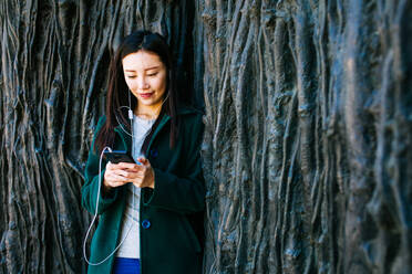 Schöne asiatische Frau in stilvollem Outfit, die Musik hört und ihr Smartphone benutzt, während sie sich an eine raue Wand mit einem Relief aus Baumstämmen lehnt - ADSF10193