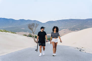 Smiling multi-ethnic couple walking on road amidst sand dunes against clear sky - OCMF01607