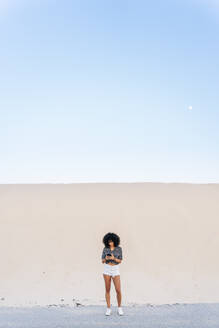 Junge Frau benutzt ein Mobiltelefon, während sie auf der Straße vor einer Sanddüne steht - OCMF01608