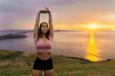 Young woman stretching arms while standing against sea during sunset - MGOF04358