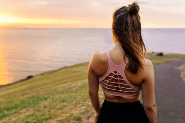 Junge Sportlerin schaut bei Sonnenuntergang aufs Meer - MGOF04359