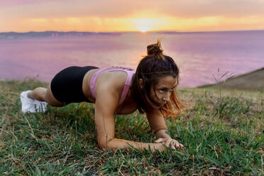 Entschlossene junge aktive Frau, die während des Sonnenuntergangs die Plankenstellung im Gras übt - MGOF04363