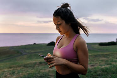 Young sportswoman using smart phone while standing against sky during sunset - MGOF04367