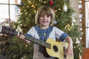 Lächelnder 4 Jahre alter Junge spielt Gitarre mit Weihnachtsbaum im Hintergrund - MINF15012