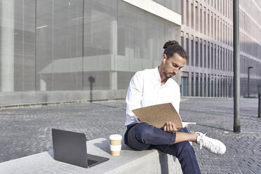Businessman reading file while sitting on block in city - VEGF02643