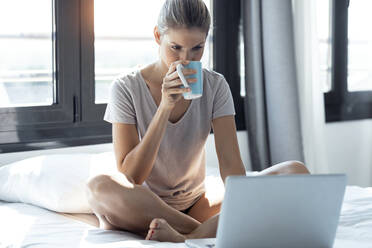 Blonde Frau sitzt auf dem Bett, benutzt einen Laptop und trinkt Kaffee - JSRF00984