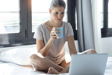 Blonde Frau sitzt auf dem Bett, benutzt einen Laptop und trinkt Kaffee - JSRF00985