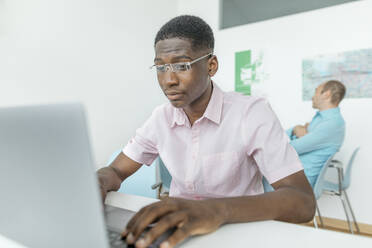 Ein Mitarbeiter arbeitet an einem Laptop, während ein Kollege im Hintergrund am Arbeitsplatz sitzt - KMKF01454
