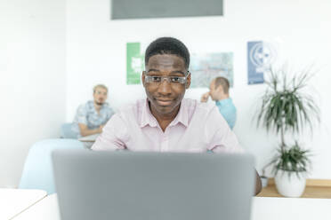 Business person working on laptop while colleagues sitting in background at workplace - KMKF01455