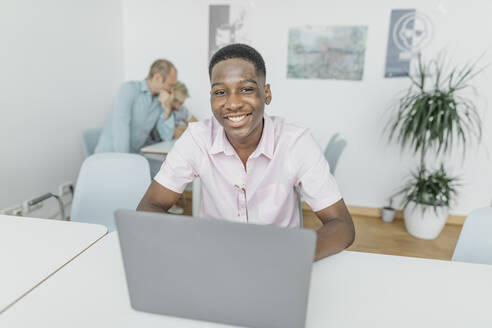 Lächelnder Geschäftsmann, der an einem Laptop arbeitet, während seine Kollegen im Hintergrund am Arbeitsplatz sitzen - KMKF01457