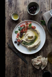 Von oben: Teller mit köstlichem Pesto-Hummus, dekoriert mit Kirschtomaten und Bohnen mit Petersilie, auf einem Holztisch neben Fladenbrot und Öl - ADSF10327