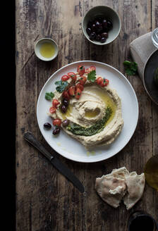 Von oben: Teller mit köstlichem Pesto-Hummus, dekoriert mit Kirschtomaten und Bohnen mit Petersilie, auf einem Holztisch neben Fladenbrot und Öl - ADSF10327