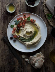 Von oben: Teller mit köstlichem Pesto-Hummus, dekoriert mit Kirschtomaten und Bohnen mit Petersilie, auf einem Holztisch neben Fladenbrot und Öl - ADSF10330