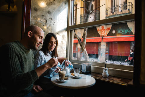 Gutaussehender Mann mit Glatze und attraktive Frau genießen Kaffee und Tee, während sie an einem kleinen Tisch in der Nähe des Fensters sitzen und sich in einem gemütlichen Café verabreden - ADSF10378