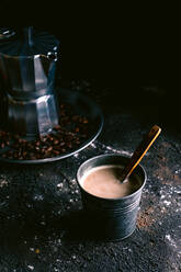 Metalltasse mit frisch gebrühtem Kaffee und Holzlöffel auf unordentlicher schwarzer Fläche neben Tablett mit Kaffeemaschine und gerösteten Körnern - ADSF10396