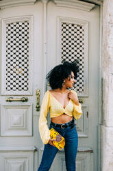 Trendy African American woman in yellow crop top and jeans standing by door and looking away - ADSF10417