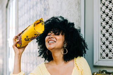 Cheerful young woman with lush curly hair and closed eyes holding fashion bag in raised hand and laughing - ADSF10418