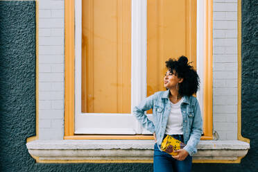 Trendy ethnischen Frau in Jeans und Jeansjacke hält Mode Handtasche und lehnt sich auf helle Fensterbank im Freien - ADSF10420