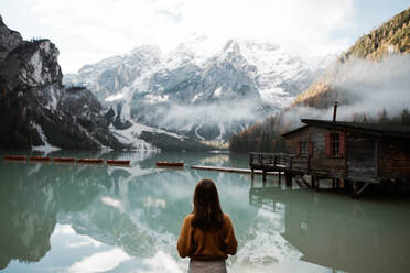 Rückenansicht einer anonymen Reisenden in Freizeitkleidung, die stehend den Blick auf ein Holzhaus am türkisfarbenen See mit nebligen Dolomitenbergen im Hintergrund in Italien genießt - ADSF10422