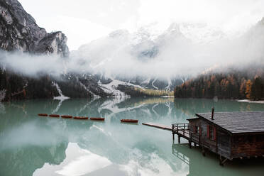 Holzhaus auf Stelzen und Boote auf nebligem See mit Spiegelung der mächtigen Dolomiten in Italien - ADSF10424