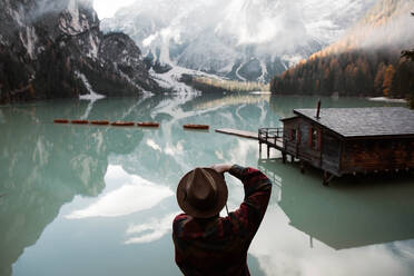 Back view of anonymous hipster man traveler in casual wear standing and delighting in view of wooden house on turquoise lake with foggy Dolomites mountains on background at Italy - ADSF10425