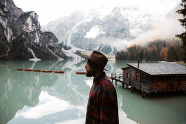 Rückenansicht eines anonymen Hipster-Mannes in Freizeitkleidung, der stehend den Blick auf ein Holzhaus am türkisfarbenen See mit nebligen Dolomitenbergen im Hintergrund in Italien genießt - ADSF10426