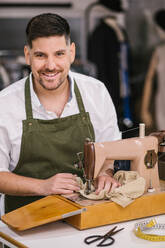 Diligent focused male tailor in apron sewing outfit details using modern sewing machine at table while creating exclusive clothes collection in contemporary work studio - ADSF10436