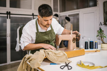 Diligent focused male tailor in apron sewing outfit details using modern sewing machine at table while creating exclusive clothes collection in contemporary work studio - ADSF10437