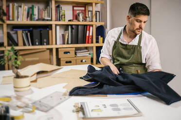 Crop young male dressmaker working with a jacket on sewing examining zipper details of future outfit while working on exclusive apparel collection at table against blurred interior of contemporary atelier - ADSF10439