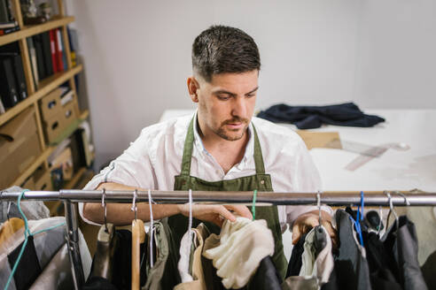 Hoher Winkel des ernsten fokussierten männlichen Schneiders in der Schürze, der Details der Kleidung überprüft, die auf Bügel auf Metallregal unter anderen trendy maßgeschneiderten Kleidern im modernen Arbeitsraum hängt - ADSF10441