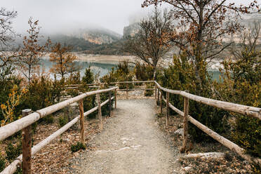 Empty curved path with wooden railing leading among trees with dry foliage on hill slope near mountain lake in foggy day in Montsec Range in Spain - ADSF10445