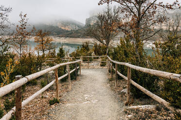 Leerer geschwungener Weg mit Holzgeländer, der zwischen Bäumen mit trockenem Laub auf einem Berghang in der Nähe eines Bergsees an einem nebligen Tag im Montsec-Gebirge in Spanien verläuft - ADSF10445