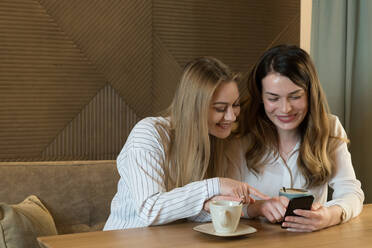 Zwei stilvolle Freundinnen mit Tassen frischem Kaffee lächelnd und Blick auf das Telefon beim Sitzen am Tisch in gemütlichen Restaurant - ADSF10508