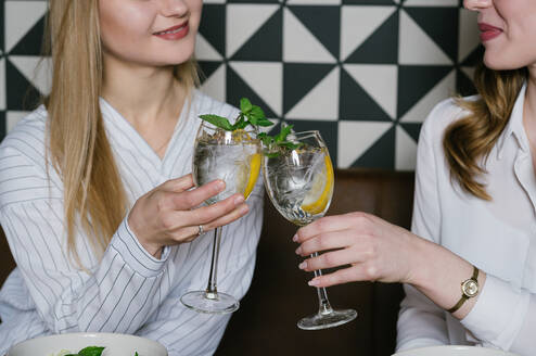 Zwei hübsche Damen lächeln und stoßen mit Gläsern von Alkohol-Cocktails an, während sie in einem gemütlichen Restaurant zu Mittag essen - ADSF10509