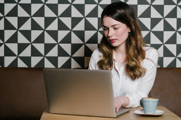 Weibliche Freiberuflerin beim Surfen auf einem modernen Laptop, während sie mit einer Tasse Kaffee in einem gemütlichen Restaurant am Tisch sitzt - ADSF10510