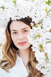 Attractive lady smiling and looking at camera while standing near tree branches blooming with white flowers - ADSF10515