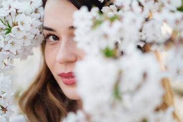 Attractive lady smiling and looking at camera while standing near tree branches blooming with white flowers - ADSF10516