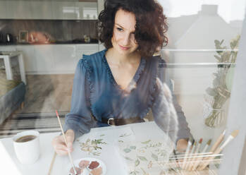 Blick durch ein Fenster auf eine elegante Frau, die mit einem Pinsel Aquarellblumen auf ein großes Blatt am Schreibtisch malt - ADSF10523