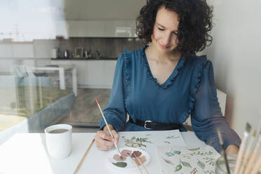 Blick durch ein Fenster auf eine elegante Frau, die mit einem Pinsel Aquarellblumen auf ein großes Blatt am Schreibtisch malt - ADSF10524