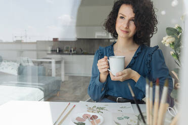 Nachdenkliche brünette Künstlerin, die mit einer Tasse Kaffee am Tisch sitzt und am Arbeitsplatz wegschaut - ADSF10526