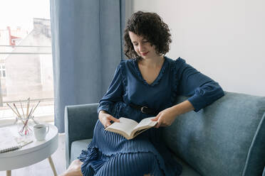Glückliche junge brünette Frau in blauem Kleid lesen Buch sitzt auf Couch am Tisch mit kreativen Accessoires Quasten und Album zu Hause - ADSF10532