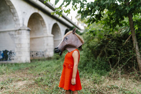 Kleines Mädchen mit Pferdekopf und rotem Kleid steht in der Natur unter einer Brücke - EGAF00645